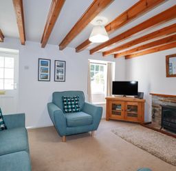 A living room with wooden beams on the ceiling blue armchairs a wooden TV stand with a television and a fireplace at Pennine in Veryan