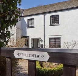A white cottage with black-framed windows and an open door behind a gate with New Mill Cottages sign at 2 New Mill Cottages in North Bovey