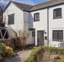 A white building with black window frames and a water wheel surrounded by bushes and a gravel path at 2 New Mill Cottages in North Bovey