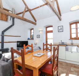 A kitchen and dining area with wooden beams on the ceiling a wooden dining table with chairs a gray sofa a black stove television and windows at The Yarde Annexe in Pen Elm near Taunton