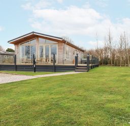 A wooden cabin with large glass windows and a deck surrounded by glass railings on a grassy lawn at 6 Meadow Retreat in Dobwalls