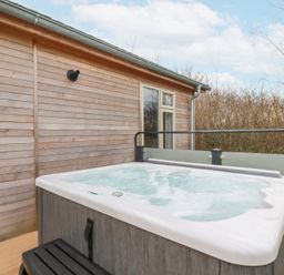 An outdoor hot tub on a wooden deck next to a wooden house wall with trees in the background at 6 Meadow Retreat in Dobwalls