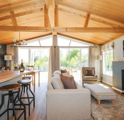 A living room with beige sofa armchair plaid ottoman wood stove and kitchen bar stools at 17 Meadow Retreat in Dobwalls