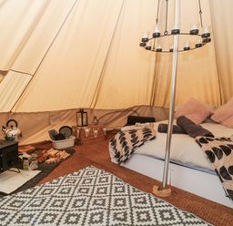 Interior of a tent with a bed, wood stove, firewood, kettle, cushions, blankets, and a hanging candle holder at Bell tent 4 in Camerton near Timsbury