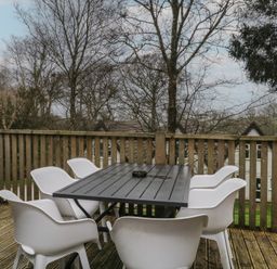 A wooden outdoor deck with a black table and six white chairs surrounded by trees at Valley Lodge 38 in St Ann's Chapel Cornwall