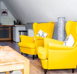 Two yellow armchairs with bicycle print cushions in a living area next to a wooden coffee table and kitchen in Valley Lodge 40 St Ann's Chapel Cornwall
