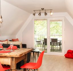 A dining area with a wooden table and red chairs next to a living space with a couch and a small table by glass doors leading to a balcony at Valley Lodge 3 in St Ann's Chapel Cornwall