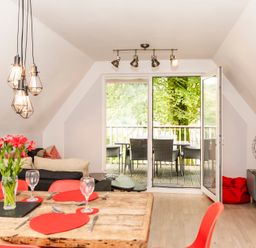 A dining and living room with a wooden table set with red heart-shaped placemats and wine glasses a black sofa and a tv unit with a television and open doors leading to a balcony with outdoor chairs at Valley Lodge 3 in St Ann's Chapel Cornwall