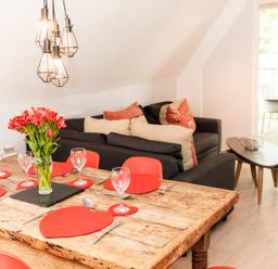 A dining table set with red heart-shaped placemats and a vase of flowers next to a living area with a dark sofa and cushions at Valley Lodge 3 St Ann's Chapel Cornwall