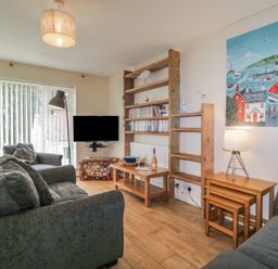 A living room with two grey sofas a wooden bookshelf a wall-mounted television and a painting of a seaside village at 26 Manorcombe in St Ann's Chapel Cornwall