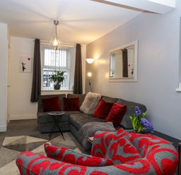 A living room with a grey sectional sofa with red and grey cushions a red patterned armchair a geometric rug and a window with dark curtains at Colliers Cottage in Cinderford
