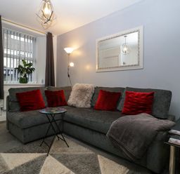 A living room with a gray sectional sofa red pillows a floor lamp a decorative mirror and a small round table at Colliers Cottage in Cinderford