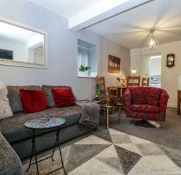A living room with a gray sectional sofa with red pillows a red patterned armchair a small round table a geometric patterned rug wooden chairs and a dining table with a lamp and wall clock at Colliers Cottage in Cinderford