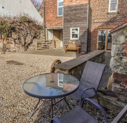 An outdoor patio with gravel ground a round glass table two chairs a stone and brick wall and a wooden bench at Seafield Granary in Alnmouth
