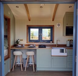 A kitchenette with two stools plates mugs and a sink under a window at Fir Tree Shepherd's Hut in Llanon