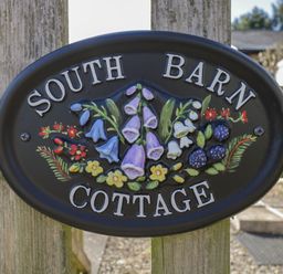 A black oval sign with flowers and berries reading South Barn Cottage on a wooden fence at South Barn Cottage in Hatford near Stanford In The Vale