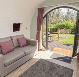 A seating area with a sofa and purple pillows next to a large arched glass door opening to a deck and garden at The Rowan Pod in Cowbridge