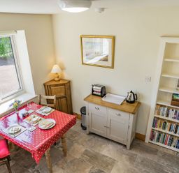 A dining area with a table set for two near a window a cabinet with a toaster and kettle and a bookshelf filled with books at Pant Y Cwtsh in Kidwelly