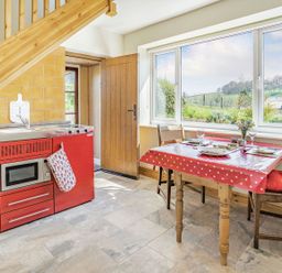 A kitchen with a red stove and microwave a wooden dining table with a red polka dot tablecloth and two chairs near a window at Pant Y Cwtsh in Kidwelly