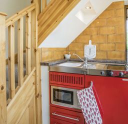 A small kitchen area with red cabinets a sink a microwave and a stove next to wooden stairs and a door to outside at Pant Y Cwtsh in Kidwelly