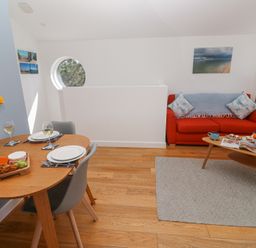 A dining area with a wooden table set for two and a living room with a red sofa and coffee table at Azure Retreat at Una in Carbis Bay near St Ives