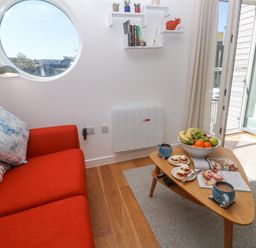 A sitting area with an orange sofa, a wooden coffee table with mugs, fruit bowl, and snacks near a round window and open door at Azure Retreat at Una in Carbis Bay near St Ives