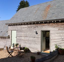 An outdoor patio with gravel floor a small table and two wooden chairs against a wooden paneled building at The Dairy at Wyke Farm Wyke near Lyme Regis