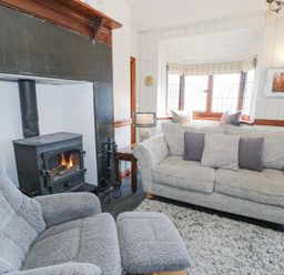 A living room with a wood-burning stove a grey armchair and sofa with cushions in front of a bay window at Bwthyn y Coediwr The Woodcutters Cottage in Tan Y Bwlch Maentwrog