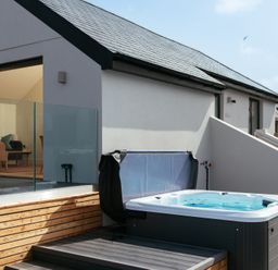 An outdoor space with a hot tub near a wooden deck and a glass railing next to a modern house at Shearwater in Mullion