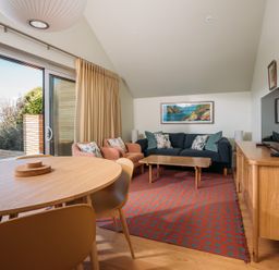 A living room with a wooden dining table and chairs a blue sofa and two armchairs a wooden cabinet with a television and a sliding glass door at Shearwater in Mullion