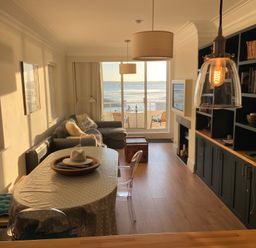 A dining area with a table and chairs next to a living room with a sofa and a bookshelf overlooking a balcony with a sea view at Flat 7 Sand Acres in Canford Cliffs