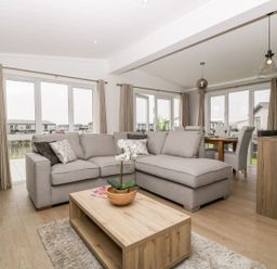 A living room with a gray sectional sofa a wooden coffee table with a plant and dining area with chairs and pendant lights at Summerdown in Brean