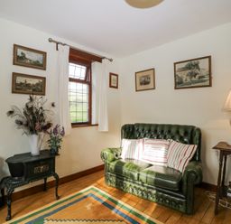 A small living room with a green leather sofa striped pillows a lamp on a wooden side table framed paintings on white walls and a decorative stove at Wringford Farm Annexe Rame near Kingsand