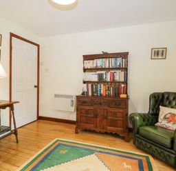 A room with a wooden bookshelf filled with books a wooden side table with a white lamp two green leather armchairs and a colorful rug on the floor at Wringford Farm Annexe Rame near Kingsand