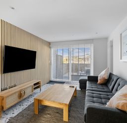 A living room with a dark gray sofa two pillows a wooden coffee table a wooden TV stand with a mounted flat screen TV and a large window with blinds at 5 Mermaid Court in Newquay