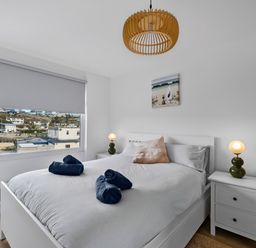 A bedroom with a bed with white bedding and blue towels on it two bedside tables with lamps and a window with a view of buildings at 5 Mermaid Court in Newquay