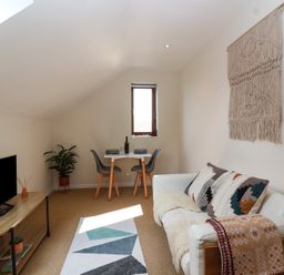 A small living room with a white sofa with patterned cushions a wooden table with a black TV a plant a dining table set for two and a wall hanging at Priory Barn Apartment in Walton near Street