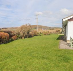 A garden with grass and bushes next to a white house with chairs on the patio at Seafield in Rhydwyn near Cemaes Bay