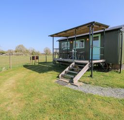 A shepherds hut with a covered porch and stairs in a grassy fenced field at Gilberry Shepherds Hut in Wall under Heywood near Church Stretton