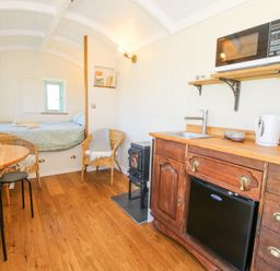 A compact living space with a bed in the background wicker chairs a small stove a wooden kitchen unit with a sink kettle microwave and a table with chairs at Gilberry Shepherds Hut Wall under Heywood near Church Stretton