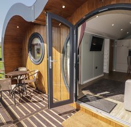 An outdoor wooden deck with a round table and four chairs leading to an open door showing a living area with a wall-mounted TV and kitchen at The Hazel Pod in Cowbridge