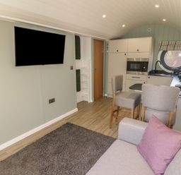 A living area with a sofa and purple pillow a wall-mounted TV a small kitchen with appliances and a round window at The Hazel Pod in Cowbridge