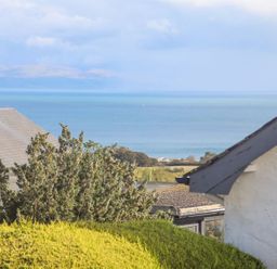 A view of a sea with rooftops, trees, and shrubs in the foreground at Trem Ynys 3 bed in Abersoch