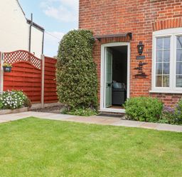 A garden with grass lawn flowers and a bush in front of a brick house with an open door at 1 Rumsby Cottages Gisleham near Kessingland