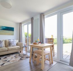 A living room with a beige sofa a wooden dining table and large glass doors at The Lookout in Perranwell near Goonhavern