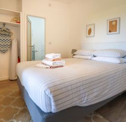 A bedroom with a bed with folded towels and pillows an open wardrobe with an ironing board and iron a bedside table and two framed pictures on the wall at The Lookout in Perranwell near Goonhavern