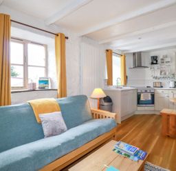A living room with a blue sofa and yellow curtains next to a kitchen with wooden dining furniture at Hollies Cottage in Perranwell near Goonhavern