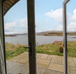 A view of a lake and hills through open glass doors with a stone patio and grass at Creags in Cullipool Isle Of Luing