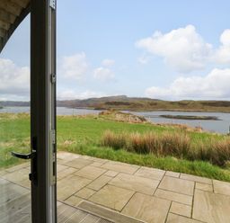 A patio door opening to a stone terrace with a grass lawn and water view at Erraid in South Cuan near Cullipool Isle Of Luing