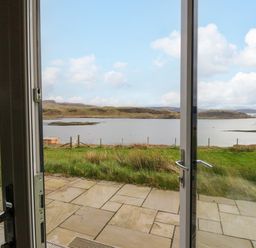 A view of a grass lawn and water with hills in the distance seen through open glass doors at Insh Cullipool Isle Of Luing